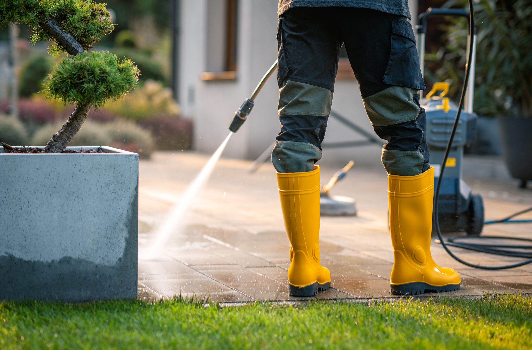 A person wearing yellow rubber boots operates a pressure washer, cleaning a patio area surrounded by greenery. The scene conveys a bright, sunny day in a residential setting.